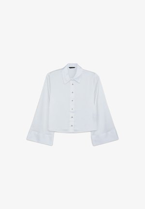 Blusa bianca a maniche lunghe, corta e con ampi polsini, chiusura a bottoni e colletto puntato, presentata su uno sfondo bianco.