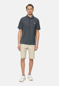 Marineblaues Poloshirt mit kurzen Ärmeln, strukturiertem Muster, Kragen und Brusttasche, kombiniert mit beigen Shorts und weißen Sneakers.