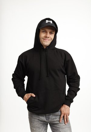 Young man wearing a black hoodie and black Under Armour cap, smiling with one hand in hoodie pocket and the other resting on jeans.