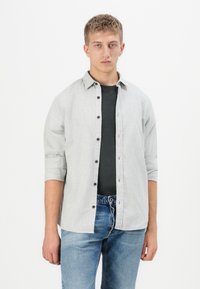 Giovane uomo con le maniche arrotolate, camicia grigia a bottoni sopra una t-shirt nera e jeans blu, in piedi su uno sfondo bianco.