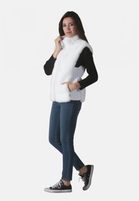 Gilet en fausse fourrure blanche avec col haut, fermeture éclair et poches latérales, porté sur un t-shirt noir à manches longues, associé à des jeans et des baskets.