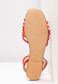 Sandale avec des brides rouges et une semelle beige, placée verticalement contre une surface d'angle blanche, montrant la semelle et le texte de la marque.