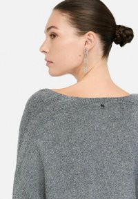 Pull en maille grise avec un large col, présentant une texture subtile. Accessoirisé avec des boucles d'oreilles en argent. Détail du logo au dos.