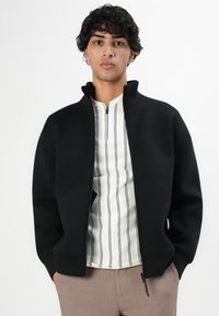 Joven con cabello rizado que lleva una chaqueta negra con cremallera sobre una camisa blanca con rayas verticales negras y pantalones beige, con las manos en los bolsillos.