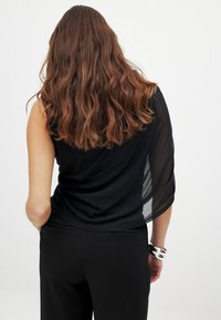 Sort top med en skulderdesign, der har et tekstureret stof og en gennemsigtig overlay langs siden. Kombineret med sorte bukser og sølv detaljer.