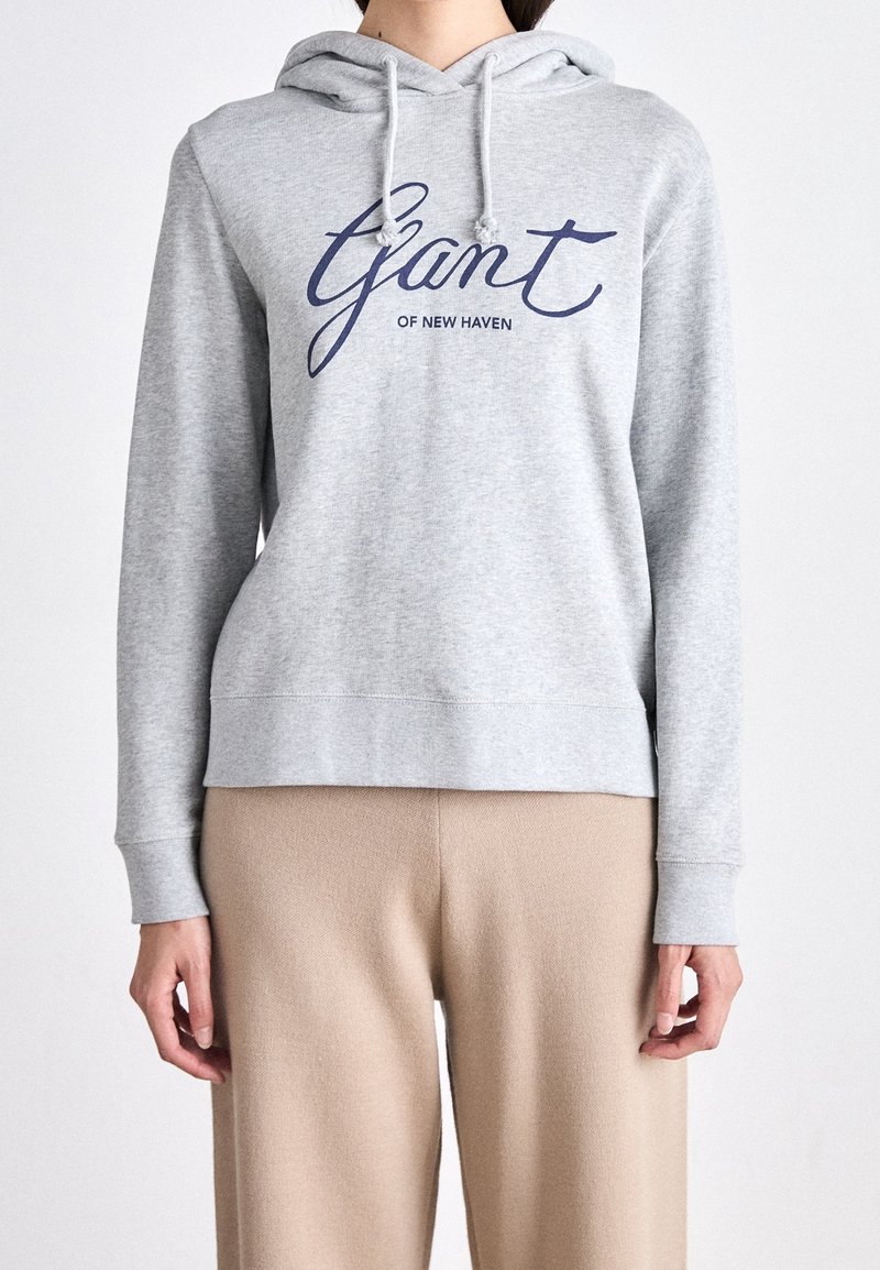 Personne portant un sweat à capuche gris clair avec le texte "Gant of New Haven" en bleu et un pantalon beige, montré du cou jusqu'au milieu de la cuisse.