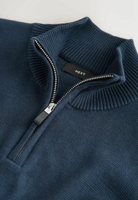 Dunkelblauer Pullover mit hohem gerippten Kragen, halbem Reißverschluss aus Metall auf der Vorderseite und glatter Strickstruktur. Etikett zeigt den Markennamen "NEXT."
