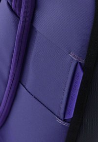 Mochila de tela morada con superficie texturizada, que cuenta con una cremallera y una etiqueta morada con la inscripción "INFORMACIÓN DEL PROPIETARIO DE LA BOLSA".