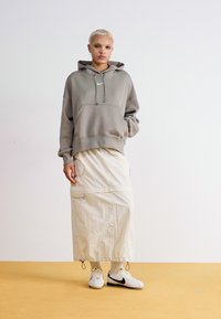 Sweatshirt cinza com capuz e bolso frontal, combinado com uma saia creme até ao joelho com bolsos de carga e ténis brancos com detalhes pretos.