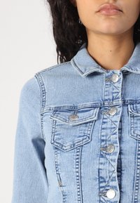 Ljusblå denimjacka med krage, tv å bröstfickor, silverknappar och sömdetaljer. Texturen är slät med en klassisk design.