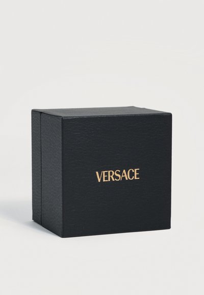 Boîte noire texturée avec logo Versace doré centré à l'avant, posée sur une surface blanche unie sur un fond clair.