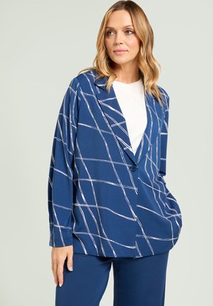 Vrouw die een blauwe blazer draagt met witte abstracte rasterlijnen over een wit topje, gecombineerd met bijpassende blauwe broek, staand tegen een lichte achtergrond.
