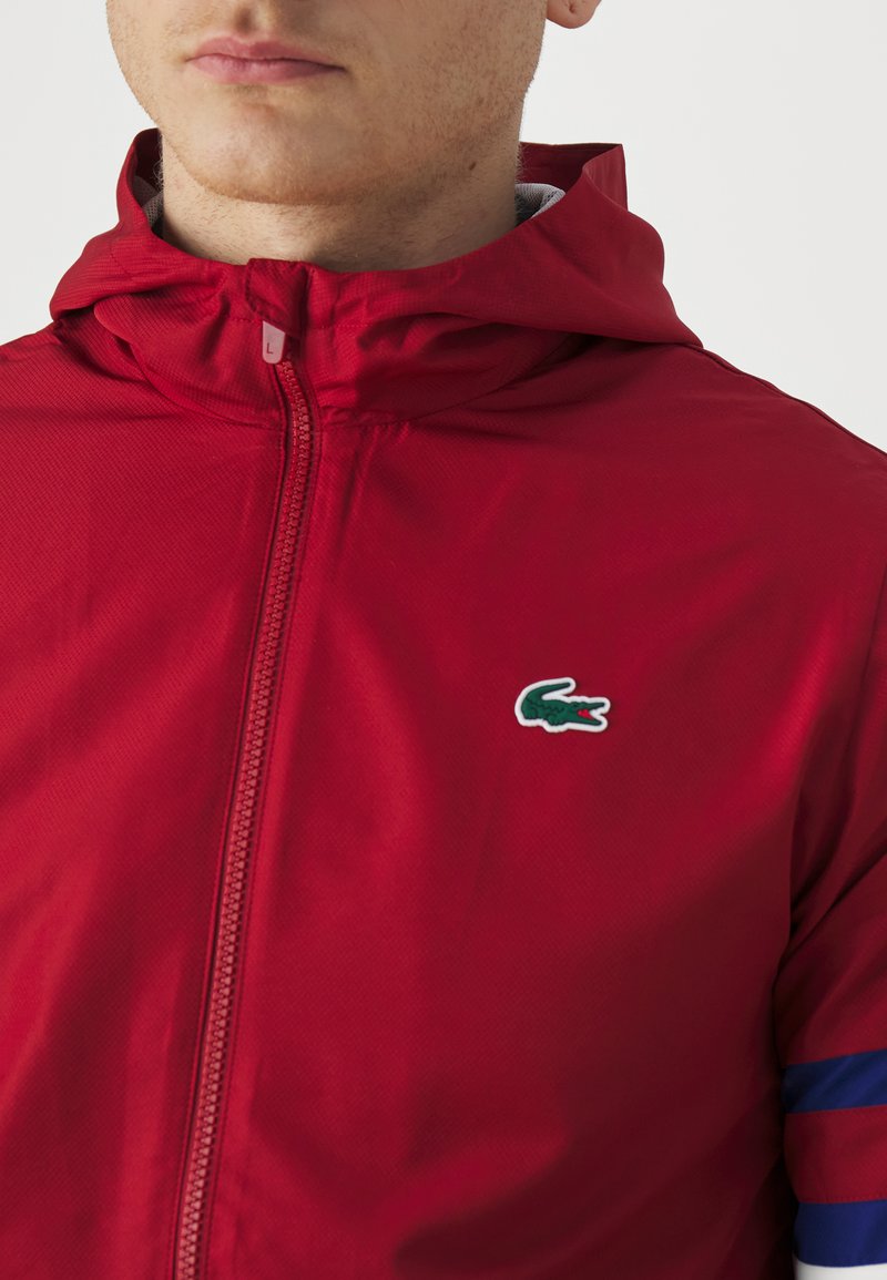 Rode ritssluitingen met capuchon, gemaakt van lichtgewicht materiaal. Heeft een klein Lacoste krokodil logo en contrasterende strepen op de mouwen.