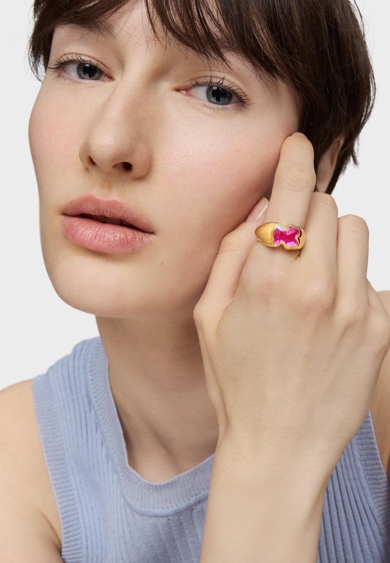 Złoty pierścionek z różowym, kokardkowym akcentem. Modelka nosi jasnoniebieską prążkowaną bluzkę, prezentując pierścionek na prawej ręce.