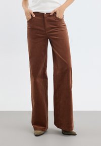 Bruna manchesterbyxor med vida ben och en texturerad yta, som har framfickor och knäppning. Tyget har vertikala ribbor.