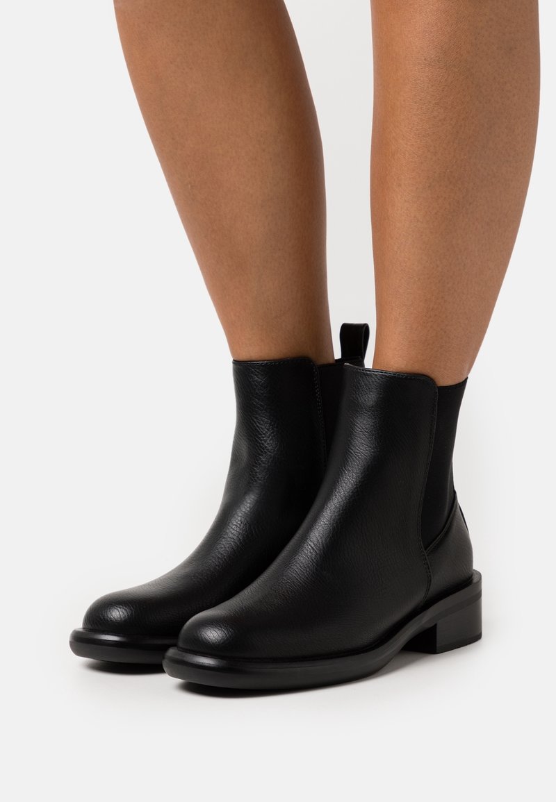 Bottines en cuir noir à talon bas portées sur des jambes nues sur fond blanc.