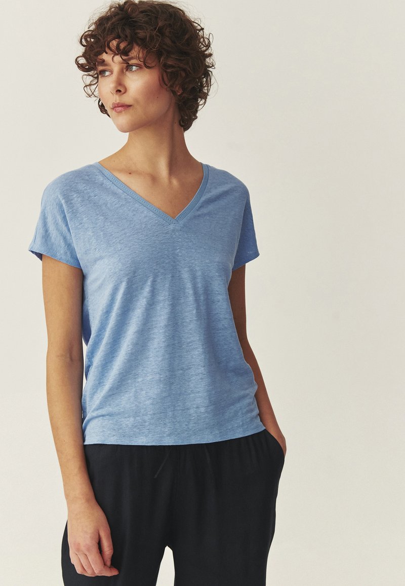 Femme aux cheveux courts et bouclés portant un t-shirt bleu clair à col en V et manches courtes, et un pantalon noir, regardant vers sa gauche avec les mains dans les poches.