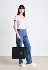 Czarna torba na ramię o gładkiej fakturze, długich uchwytach i widocznym zawieszeniu. Modelka nosi lawendowy t-shirt z dekoltem w serek i niebieskie jeansy z wysokim stanem.