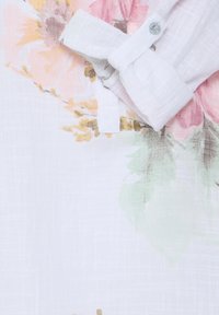 Tissu blanc avec des motifs floraux pastel en rose, jaune et vert, avec une manche retroussée et un poignet boutonné.