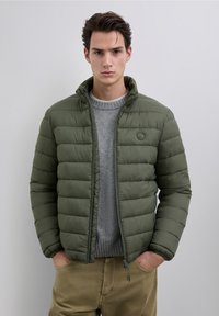 Chaqueta acolchada de color verde oliva con cierre de cremallera y cuello alto, con un diseño acolchado. Lleva puesta una sudadera gris claro.