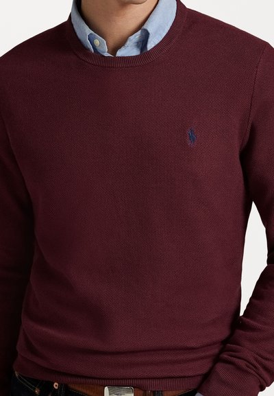 Suéter de punto burdeos con cuello redondo, tejido texturizado y un pequeño logotipo bordado en el pecho, superpuesto a una camisa de collar azul claro.