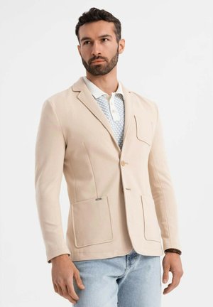 Hombre con barba vestido con un blazer beige sobre una camiseta polo blanca con estampado y jeans azul claro, de pie frente a un fondo blanco liso.