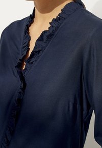 Marineblaue Bluse mit einem gerüschten Ausschnitt, weicher Textur und leicht durchscheinendem Erscheinungsbild, mit tailliertem Schnitt und minimaler Verzierung.