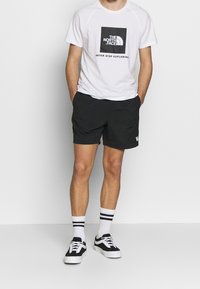 Valkoinen lyhythihainen t-paita mustalla logolla, yhdistettynä mustiin shortseihin. Mustavalkoiset lenkkarit ja valkoiset sukat mustilla raidoilla.