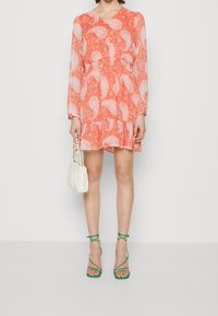 Ein pastellorange Kleid mit Paisleymuster, langen Ärmeln, einer gerafften Taille und einem Rüschensaum, kombiniert mit grünen Riemchen-Heels und einer weißen Handtasche.