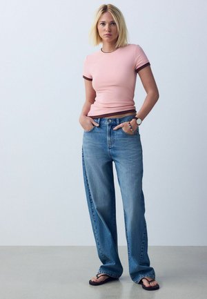 Fiatal nő áll, kezét zsebében tartva, szűk rózsaszín pólót, bő kék farmert, órát, karkötőt és fekete flip-flop papucsot viselve.