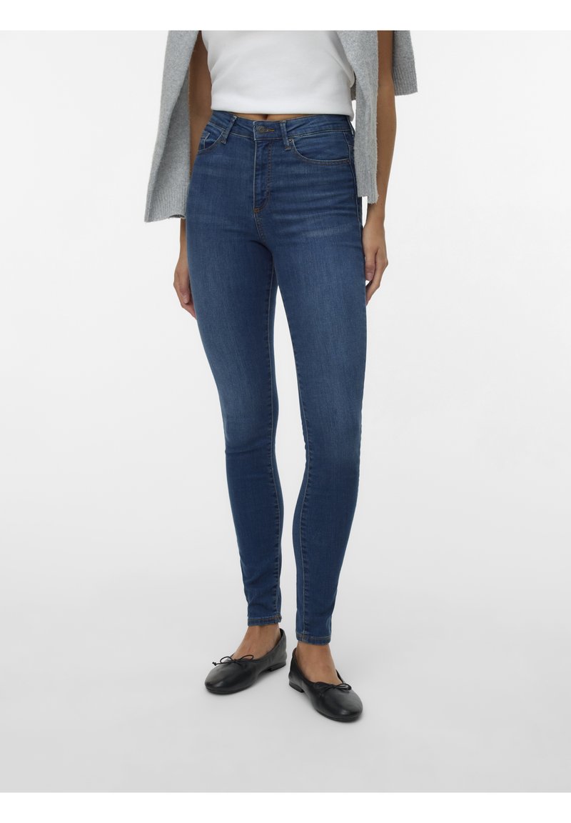 Jean skinny bleu foncé en denim stretch, avec une taille mi-haute et un design classique à cinq poches. Porté avec des ballerines noires.