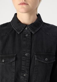 Black denim shirt featuring a spread collar, snap buttons, and two chest pockets with flaps. The fabric has a smooth, slightly textured finish.