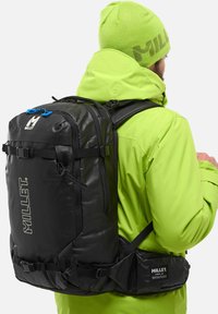 Sac à dos en millet noir avec des sangles ajustables, des accents bleus et une ceinture de hanche rembourrée, porté sur une veste vert vif.