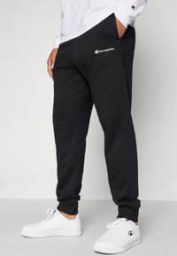Melnie sporta bikses ar sānu kabatām, elastīgiem aprocēm un izšuvu "Champion" logotipu sarkanā un baltā krāsā. Komplekts ar baltām sneakerēm.