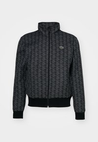 Veste noire zippée avec motif géométrique, col montant, poignets et bas côtelés, et petit logo crocodile vert sur la poitrine.
