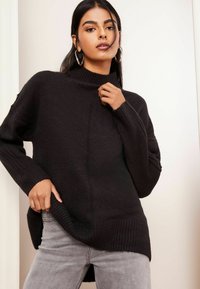 Schwarzer Rollkragenpullover aus weichem Strick, mit gerippten Bündchen und Saum, lockerer Passform und längerer Rückseite mit seitlichen Schlitzen, kombiniert mit hellgrauen Jeans.