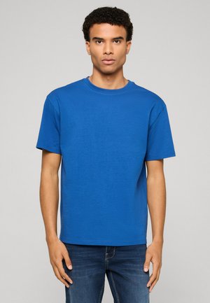 Jeune homme aux cheveux bouclés portant un t-shirt bleu uni à manches courtes et un jean bleu foncé, debout devant un fond neutre.