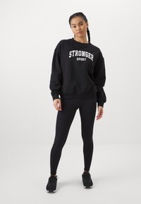 Black sweatshirt with "STRONGER SPORT" in white lettering, paired with black leggings. Features a relaxed fit and soft fabric. Black athletic shoes.