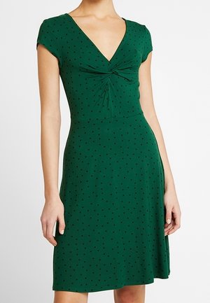 Robe verte mi-genoux à pois noirs, manches courtes, encolure en V et détail froncé torsadé au niveau de la poitrine.