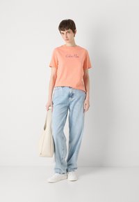 Persiko spalvos medvilninis marškinėliai su "Calvin Klein" logotipu, derinami su šviesiai mėlynomis džinsinėmis kelnėmis ir laikant kreminės spalvos petinę rankinę. Avimi baltus sportbačius.