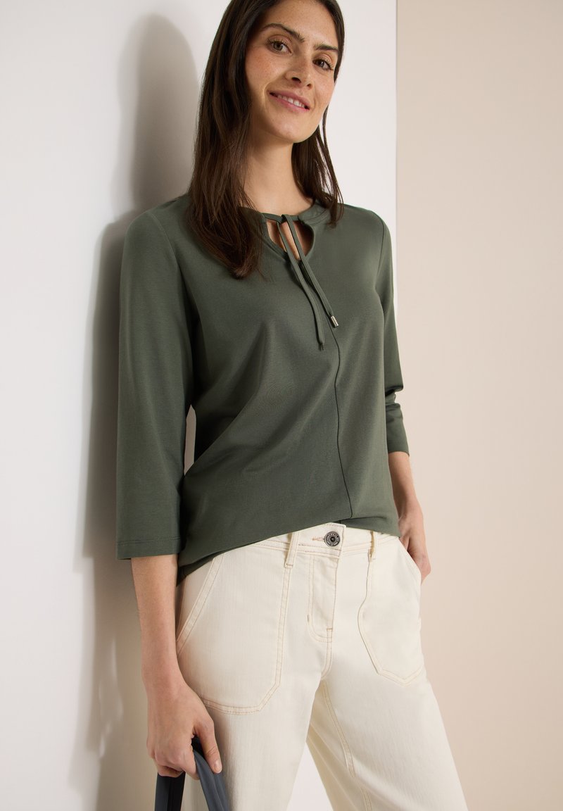 Femme portant un haut à manches longues vert olive avec un col noué et un pantalon crème taille haute, debout contre un mur bicolore beige.