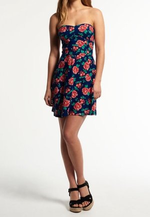 Femme portant une robe bleu marine sans bretelles avec un imprimé floral rose et des sandales à plateforme noires, posant devant un fond blanc uni.