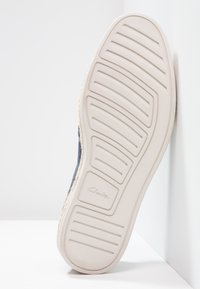 Semelle de chaussure en tissu navy avec une semelle en caoutchouc crème présentant des rainures horizontales et le logo "Clarks" embossé près du talon.