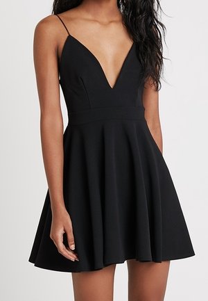 Vrouw die een mouwloze zwarte jurk draagt met spaghettibandjes en een diepe V-hals, met een getailleerde taille en een wijde rok.