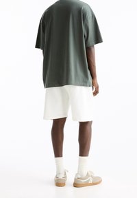 T-shirt en coton vert, coupe décontractée, short en coton blanc, et baskets beiges avec des accents blancs. Design minimaliste, tissu texturé, look décontracté.