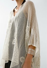 Osoba nosząca kremowy szal szydełkowy z kwiatowymi wzorami na czarnym ubraniu, uzupełniony szerokim srebrnym bransoletą na przedramieniu.