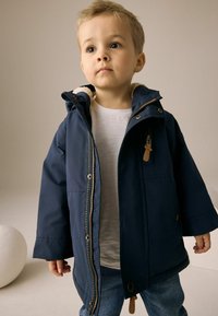 Veste pour enfant bleu marine avec capuche, fermeture éclair et poches latérales. Doublée en polaire de couleur claire, elle présente des boutons-pression et des accents en cuir.
