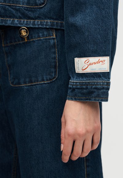 Κοντινό πλάνο χεριού δίπλα σε μανίκι τζιν μπουφάν με λευκή ετικέτα που γράφει "Sandro 1984 Summer Vibration."