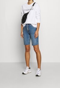 Person som bär en vit Calvin Klein sweatshirt, blå jeansshorts med knappar, vita sneakers och en svart crossover-väska framför en neutral bakgrund.