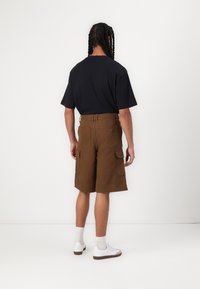 Bruna cargo-shorts med flera fickor, tillsammans med en svart t-shirt. Modell som bär vita sneakers med beige strumpor. Enfärgad bakgrund.